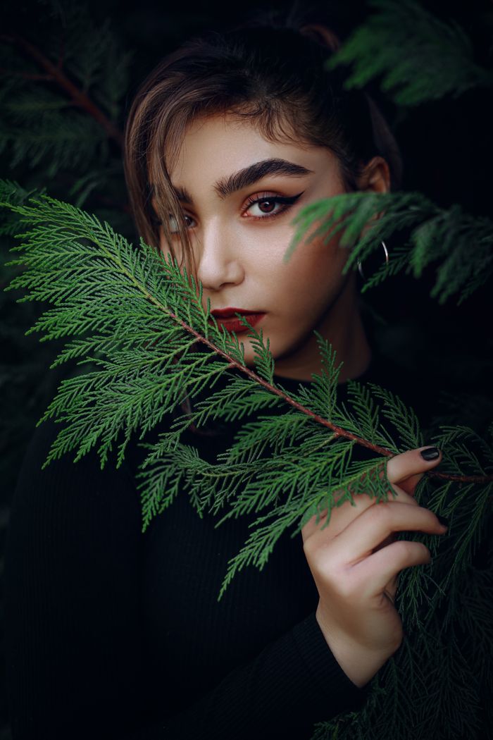 Elegant portrait of a woman with bold makeup, partially hidden behind vibrant green leaves.